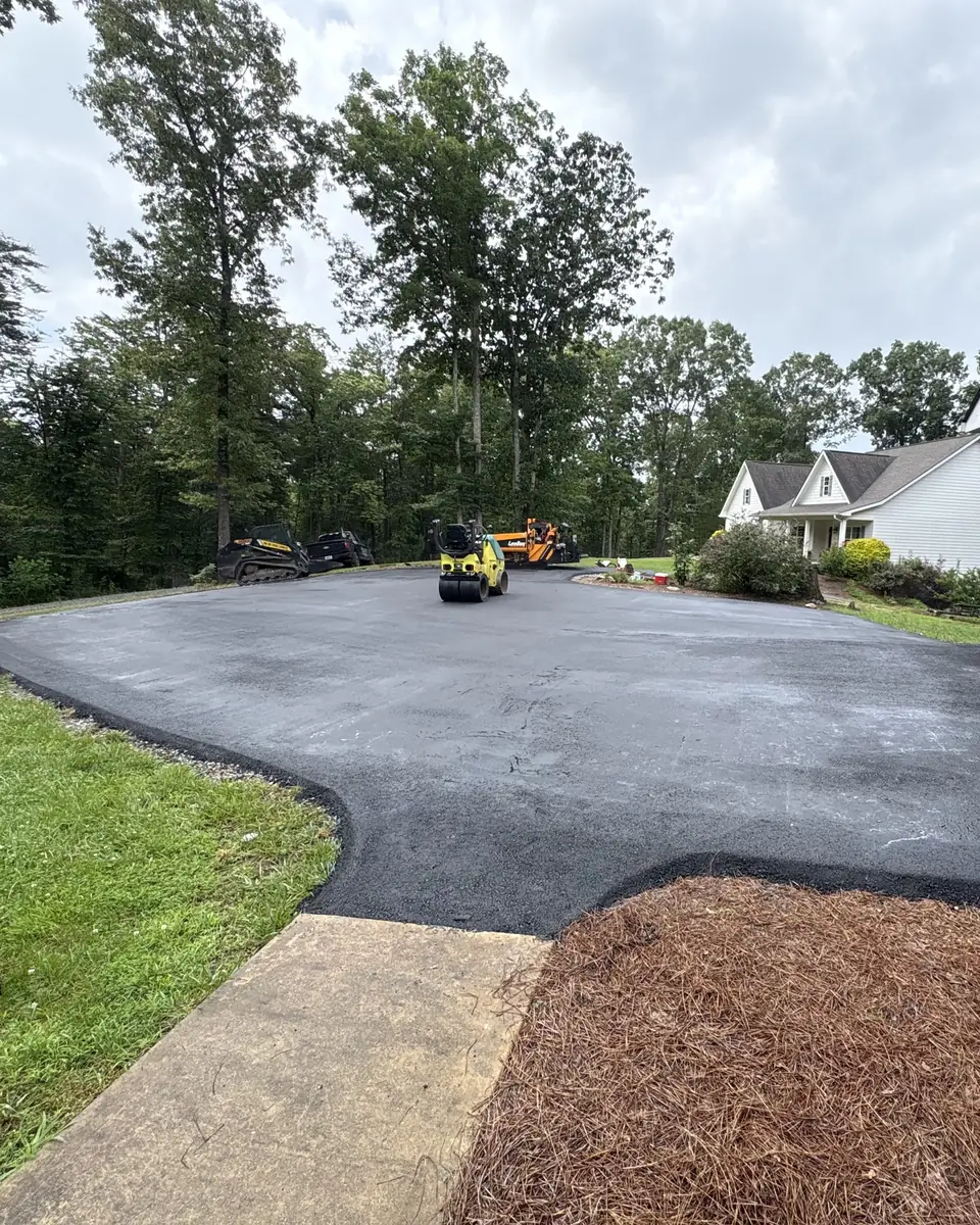 Wide new asphalt driveway sweeping toward a two-car garage, roller and paver at the far end
