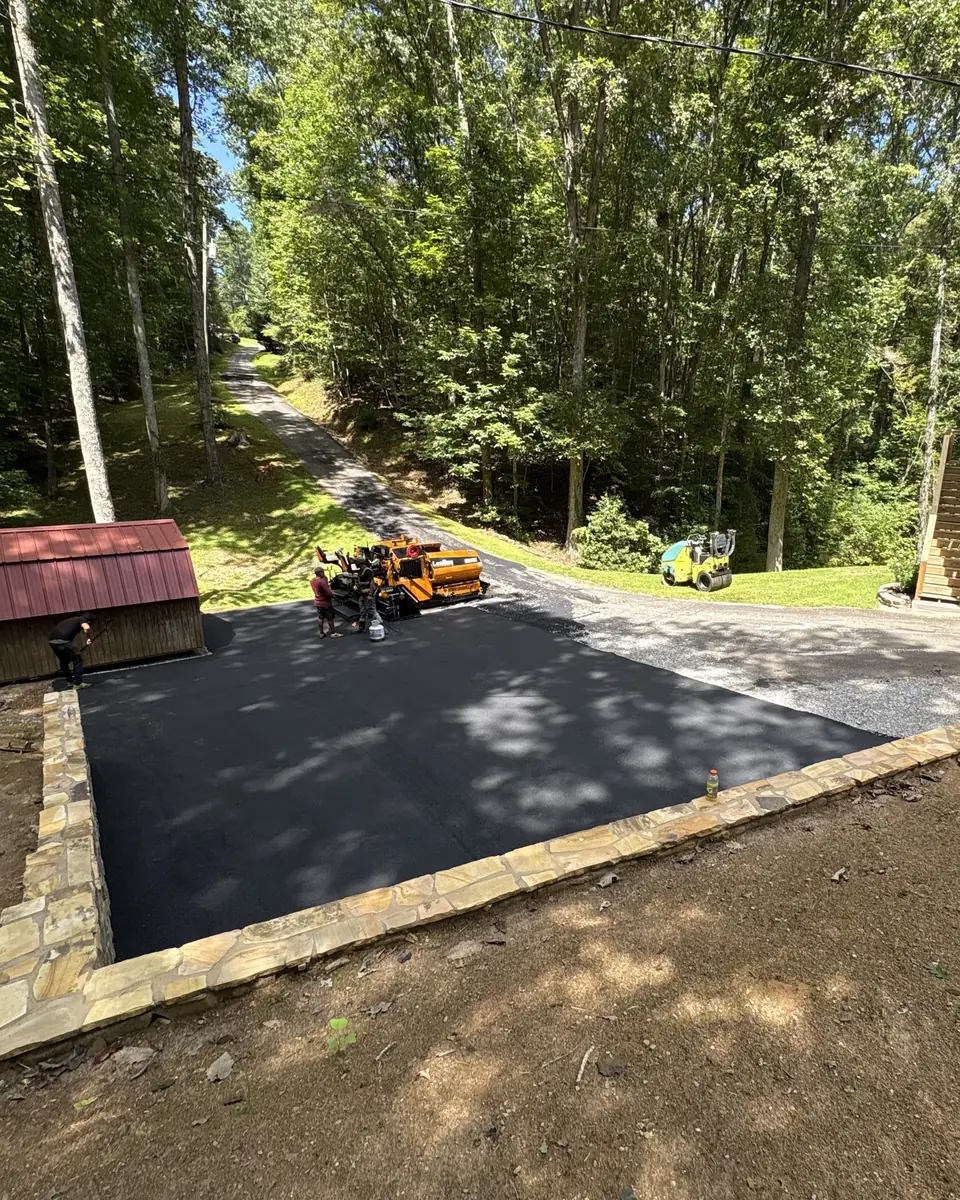 Fresh asphalt parking pad framed by a stacked-stone border, with the paver finishing the drive behind it